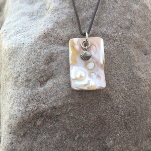 May include: A rectangular pendant necklace with a brown leather cord. The pendant is a cream and tan color with a small silver seashell charm. The necklace is displayed on a textured, gray surface.