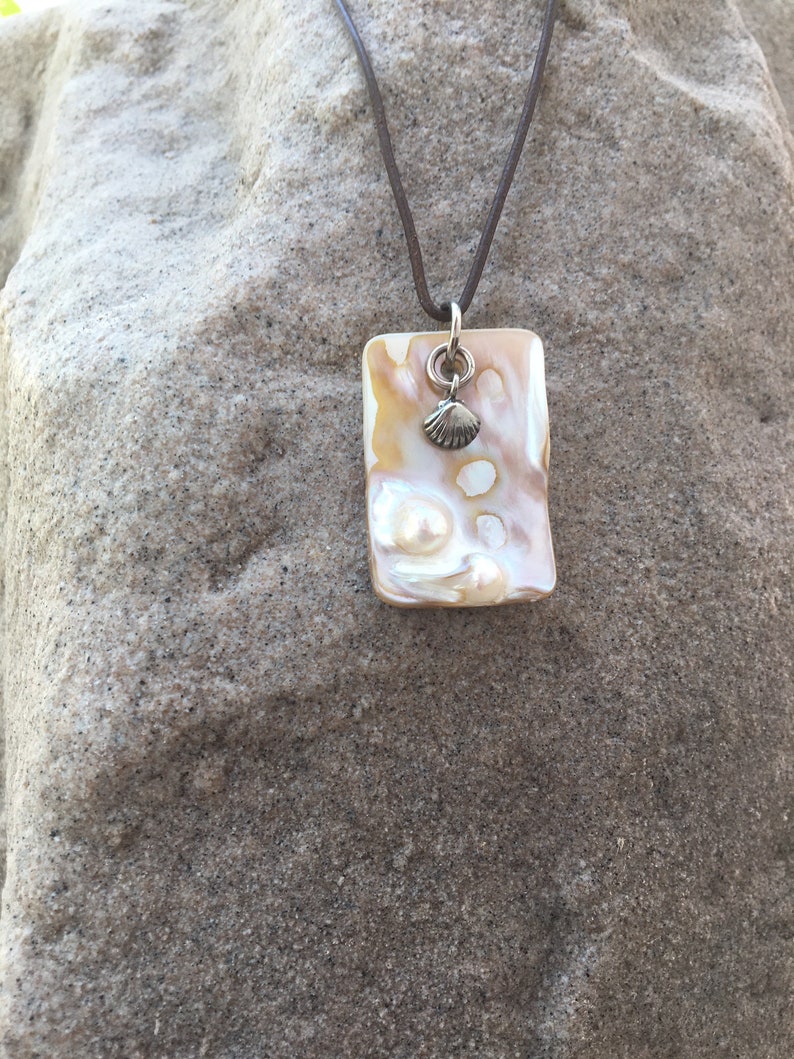 Op de afbeelding: Een rechthoekige hangerketting met een bruin leren koord. De hanger is cr&egrave;me en bruin met een kleine zilveren schelpenbedel. De ketting wordt gepresenteerd op een getextureerd, grijs oppervlak.