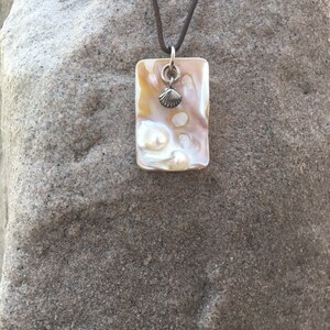 Op de afbeelding: Een rechthoekige hangerketting met een parelmoer, iriserend oppervlak. De hanger is versierd met een kleine zilveren schelpenbedel en hangt aan een bruin koord. De ketting wordt gepresenteerd tegen een getextureerde, grijze achtergrond.