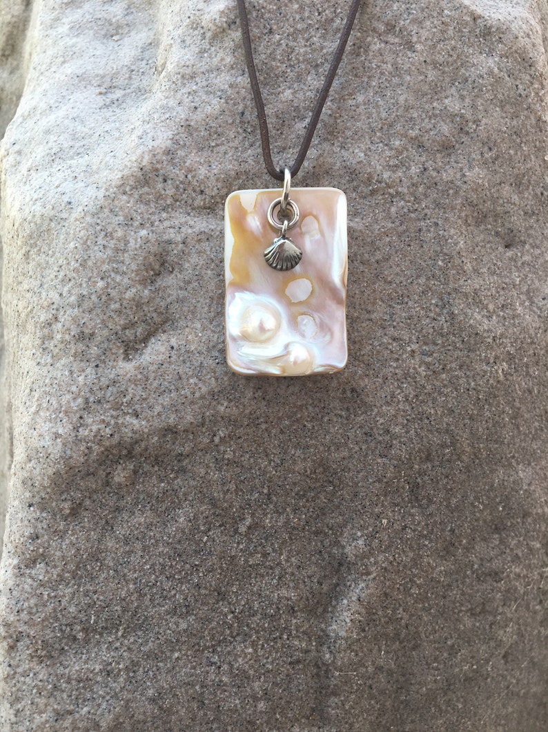 Op de afbeelding: Een rechthoekige hangerketting met een parelmoer, iriserend oppervlak. De hanger is versierd met een kleine zilveren schelpenbedel en hangt aan een bruin koord. De ketting wordt gepresenteerd tegen een getextureerde, grijze achtergrond.