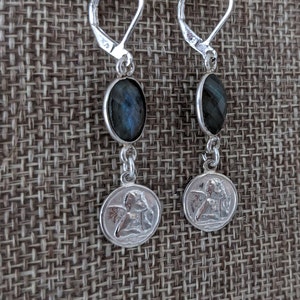 Peut inclure: Paire de boucles d'oreilles en argent avec fermoir &agrave; levier. Chaque boucle d'oreille pr&eacute;sente une pierre ovale bleu fonc&eacute; et un petit pendentif rond avec un motif d'ange. Les boucles d'oreilles sont pr&eacute;sent&eacute;es sur un fond textur&eacute; de ton neutre.