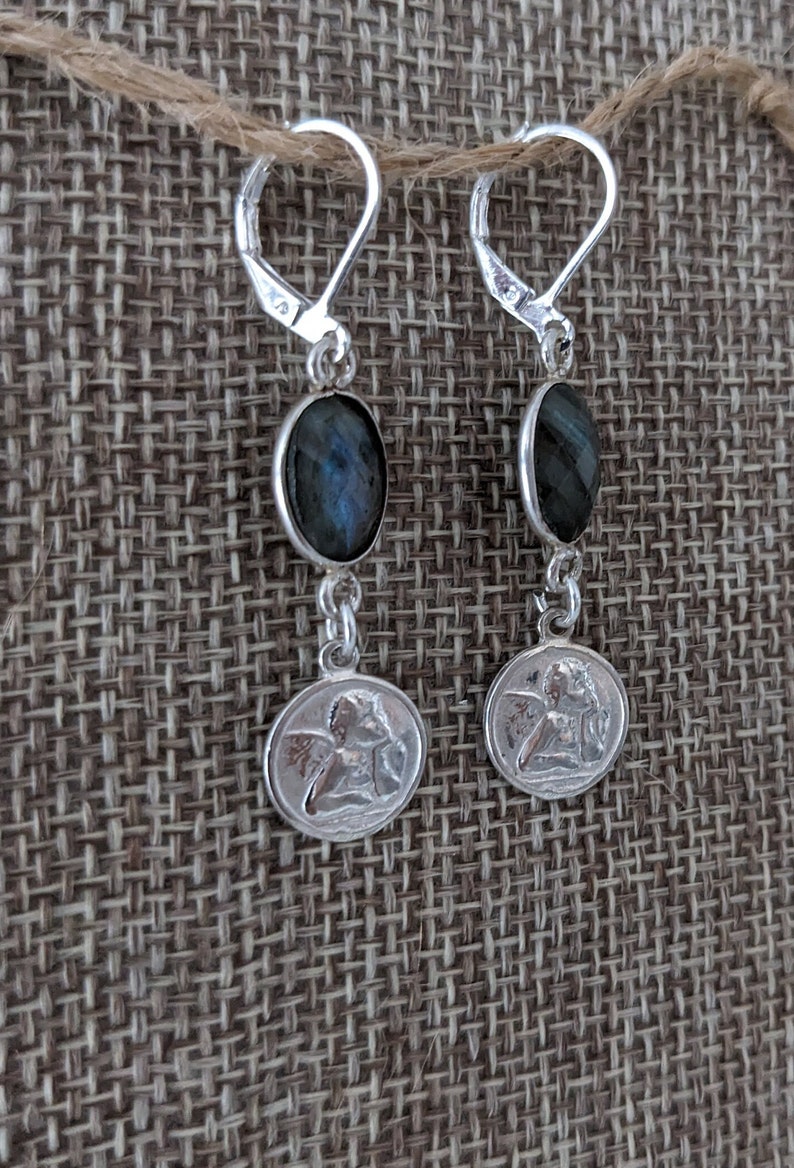 Peut inclure: Paire de boucles d'oreilles en argent avec fermoir &agrave; levier. Chaque boucle d'oreille pr&eacute;sente une pierre ovale bleu fonc&eacute; et un petit pendentif rond avec un motif d'ange. Les boucles d'oreilles sont pr&eacute;sent&eacute;es sur un fond textur&eacute; de ton neutre.