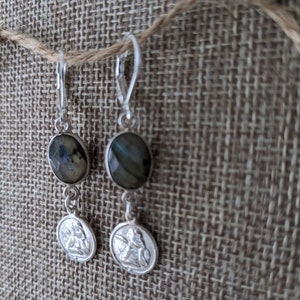 Peut inclure: Boucles d'oreilles en argent avec des pierres ovales et iris&eacute;es et de petits pendentifs ronds avec un relief d&eacute;taill&eacute; d'une figure. Les boucles d'oreilles sont suspendues &agrave; des fermoirs &agrave; levier. Le fond est un tissu textur&eacute; de couleur neutre.
