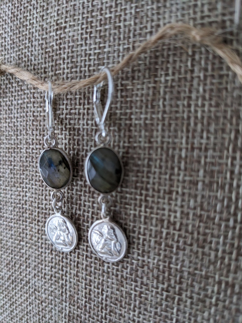 Peut inclure: Boucles d'oreilles en argent avec des pierres ovales et iris&eacute;es et de petits pendentifs ronds avec un relief d&eacute;taill&eacute; d'une figure. Les boucles d'oreilles sont suspendues &agrave; des fermoirs &agrave; levier. Le fond est un tissu textur&eacute; de couleur neutre.
