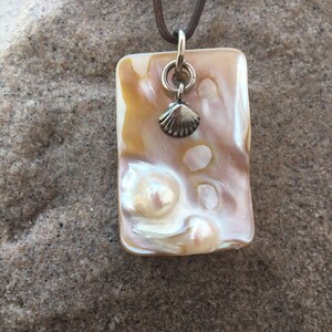 Op de afbeelding: Een rechthoekige hangerketting met een parelmoer, iriserend oppervlak in tinten cr&egrave;me, goud en roze. Een kleine zilveren schelpenbedel hangt onder een zilveren ring. De hanger hangt aan een bruin leren koord.