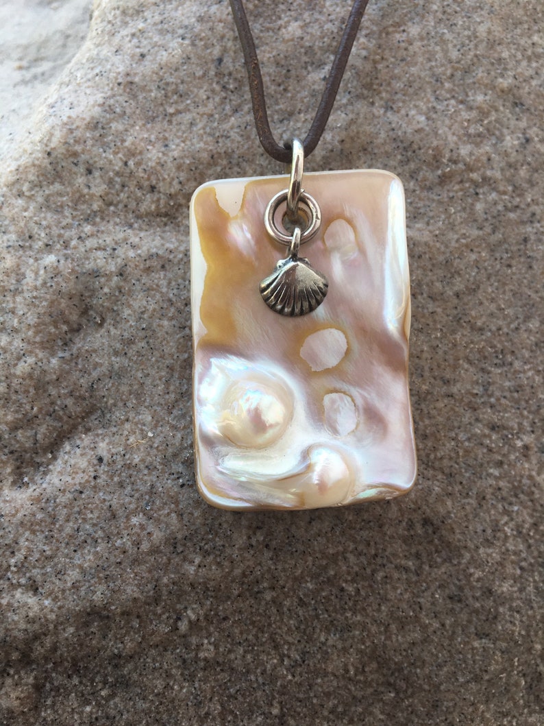 Op de afbeelding: Een rechthoekige hangerketting met een parelmoer, iriserend oppervlak in tinten cr&egrave;me, goud en roze. Een kleine zilveren schelpenbedel hangt onder een zilveren ring. De hanger hangt aan een bruin leren koord.