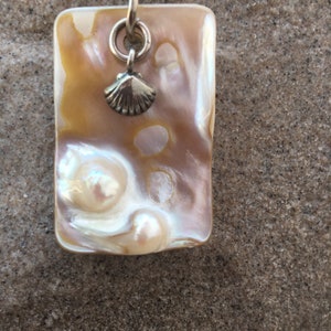 Op de afbeelding: Een rechthoekige hangerketting met een parelmoer, iriserend oppervlak in tinten cr&egrave;me, goud en roze. Een kleine zilveren schelpenbedel hangt onder een zilveren ring. De hanger is bevestigd aan een bruin leren koord.