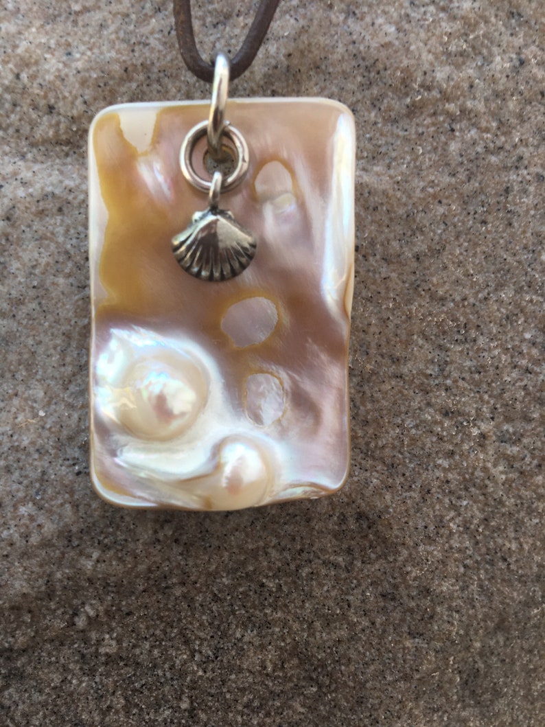 Op de afbeelding: Een rechthoekige hangerketting met een parelmoer, iriserend oppervlak in tinten cr&egrave;me, goud en roze. Een kleine zilveren schelpenbedel hangt onder een zilveren ring. De hanger is bevestigd aan een bruin leren koord.