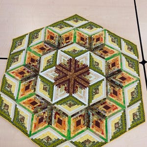 Op de afbeelding: Een zeshoekige quilt met een blokhutpatroon in tinten groen, bruin, geel en oranje. De quilt is gemaakt van stofresten en heeft een gele rand.