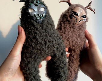 May include: Two plush, whimsical creatures with furry bodies and antler-like horns. One is dark gray with a mask adorned with leaves, the other is brown with a mask. The creatures are held up against a plain background.