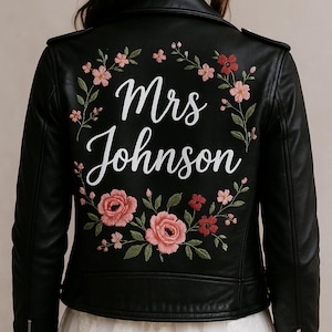 Könnte beinhalten: Schwarze Lederjacke mit einem floralen Stickmuster auf der Rückseite. Die Stickerei zeigt rosa und rote Blumen und grüne Blätter. Der Text "Mrs Johnson" ist in Weiß auf der Rückseite der Jacke gestickt.
