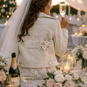 May include: Cream-colored leather jacket with floral embroidery and the text "Mrs. Taylor 06.18.2022". The jacket is worn by a person holding a champagne flute. The scene includes a table with flowers, candles, and a champagne bottle, suggesting a wedding celebration.
