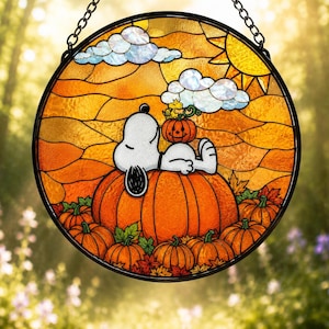 May include: A circular stained glass suncatcher featuring a white Snoopy character resting on a large orange pumpkin. A small yellow Woodstock sits on a smaller pumpkin above. The background displays an orange sky, sun, and clouds.