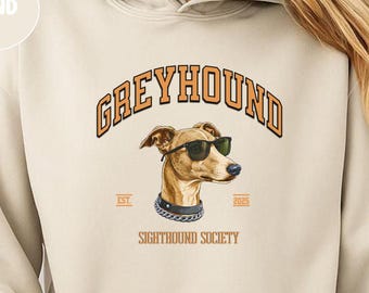 May include: Sand-colored hoodie with a front graphic of a greyhound wearing sunglasses. The text above the dog reads "GREYHOUND" in orange, with "EST. 2023" below. The hoodie has a kangaroo pocket and a drawstring hood.