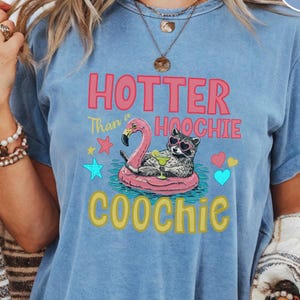 May include: A blue-jean colored t-shirt featuring a graphic of a raccoon wearing heart-shaped sunglasses while lounging on a pink flamingo pool float. The text reads HOTTER Than a HOOCHIE COOCHIE, accompanied by colorful stars and hearts.