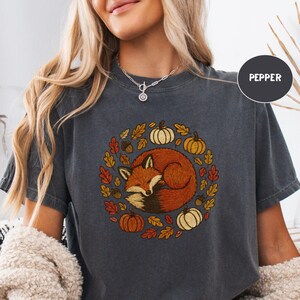 May include: Dark gray t-shirt featuring a circular design with a sleeping fox surrounded by pumpkins and autumn leaves. The word "PEPPER" is printed on the shirt. The shirt is made of a soft material.