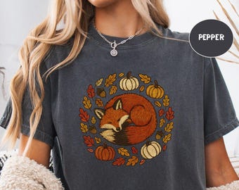 May include: Dark gray t-shirt featuring a circular design with a sleeping fox surrounded by pumpkins and autumn leaves. The word "PEPPER" is printed on the shirt. The shirt is made of a soft material.