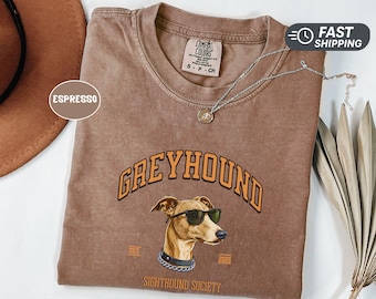 May include: Espresso-colored t-shirt featuring a greyhound wearing sunglasses and a chain collar. The word "GREYHOUND" is arched above the dog's head in orange lettering. The shirt also has the text "SIGHTHOUND SOCIETY" and the years "EST 2025".