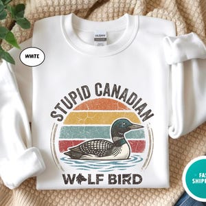 May include: White sweatshirt with a graphic design featuring a loon in water, a retro sunset, and the text "STUPID CANADIAN WOLF BIRD". The sweatshirt has long sleeves and a crew neck.