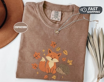 May include: Espresso-colored t-shirt with a graphic of a fox and hedgehog surrounded by autumn leaves. The shirt has a crew neck and short sleeves. A silver necklace with a pendant is draped across the shirt.