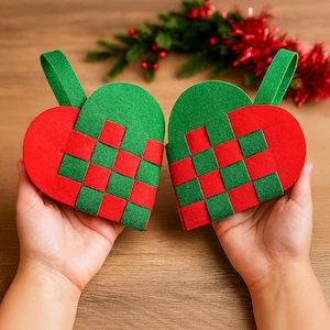 May include: Two heart-shaped Christmas ornaments. Each ornament is red and green, with a woven pattern. The top of each heart has a green loop for hanging. The ornaments are held in hands, with a wooden background.