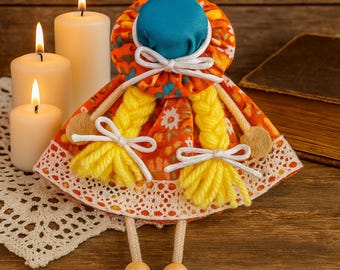 May include: A handmade doll with a blue cap, orange floral dress, and yellow yarn braids. The doll has wooden ball feet and is decorated with white bows. The doll is on a wooden surface with lit candles and a vintage book in the background.