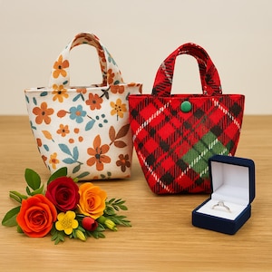 May include: Two small fabric tote bags. One is white with a floral pattern in orange, blue, and brown. The other is red plaid with a green accent and a green button closure. A blue ring box with a diamond ring is next to the red bag.