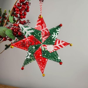 Puede incluir: Un adorno de estrella navideña hecho a mano. La estrella está hecha de tela roja y verde con diseños de copos de nieve. Se adjuntan campanas doradas y rojas a las puntas de la estrella. Se utiliza una cuerda roja y blanca para colgarla.