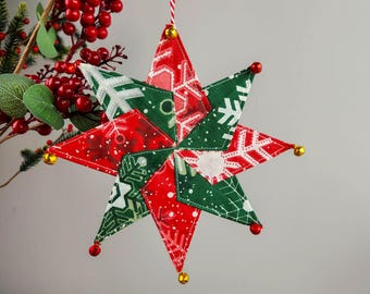 May include: A handmade Christmas star ornament. The star is made of red and green fabric with snowflake patterns. Gold and red bells are attached to the points of the star. A red and white string is used for hanging.