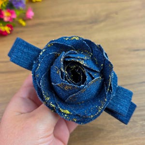 May include: A denim blue fabric rose with gold glitter accents. The flower is attached to a matching denim band. The petals are layered to create a realistic rose shape. The accessory is suitable for hair or clothing.