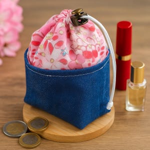 May include: A small, blue denim pouch with a pink floral drawstring bag inside. The drawstring is white, and the clasp is bronze. A red lipstick and a small perfume bottle are next to the pouch. The pouch sits on a small wooden stand.