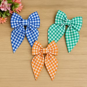 May include: Three gingham fabric hair bows in blue, green, and orange. The bows are arranged on a light brown wooden surface. Each bow has a classic bow shape with long, pointed tails. The gingham pattern is a classic checkered design.