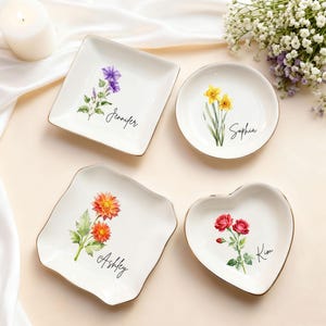 Könnte beinhalten: Vier weiße Keramikschalen mit goldenen Rändern, jede mit einem anderen Blumendesign und einem Namen. Die Formen umfassen ein Quadrat, einen Kreis, ein geschwungenes Quadrat und ein Herz. Blumen: lila, gelb, orange und rot.