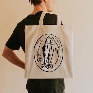 May include: A white canvas tote bag with a black illustration of two fish on a plate. The fish are drawn in a simple, stylized manner.