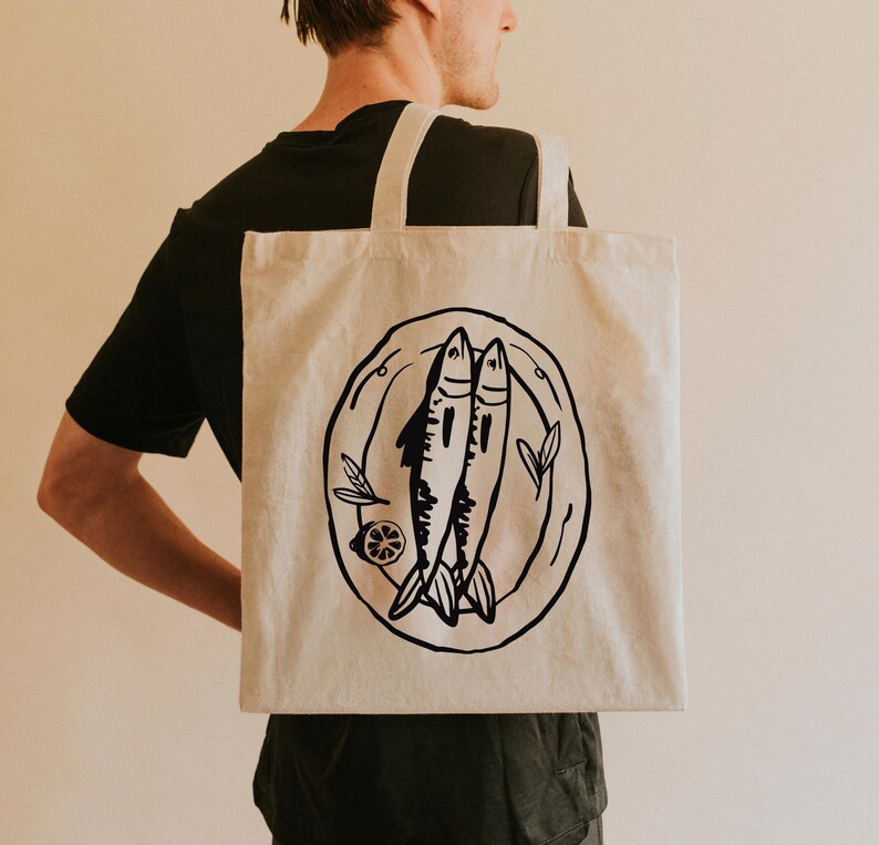May include: A white canvas tote bag with a black illustration of two fish on a plate. The fish are drawn in a simple, stylized manner.