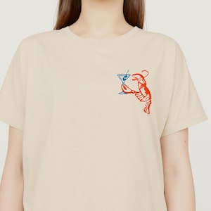 Peut inclure: T-shirt beige avec un motif rouge et bleu d'un homard tenant un verre &agrave; martini. Le homard est dessin&eacute; dans un style fantaisiste, avec le verre &agrave; martini en bleu et le homard en rouge.