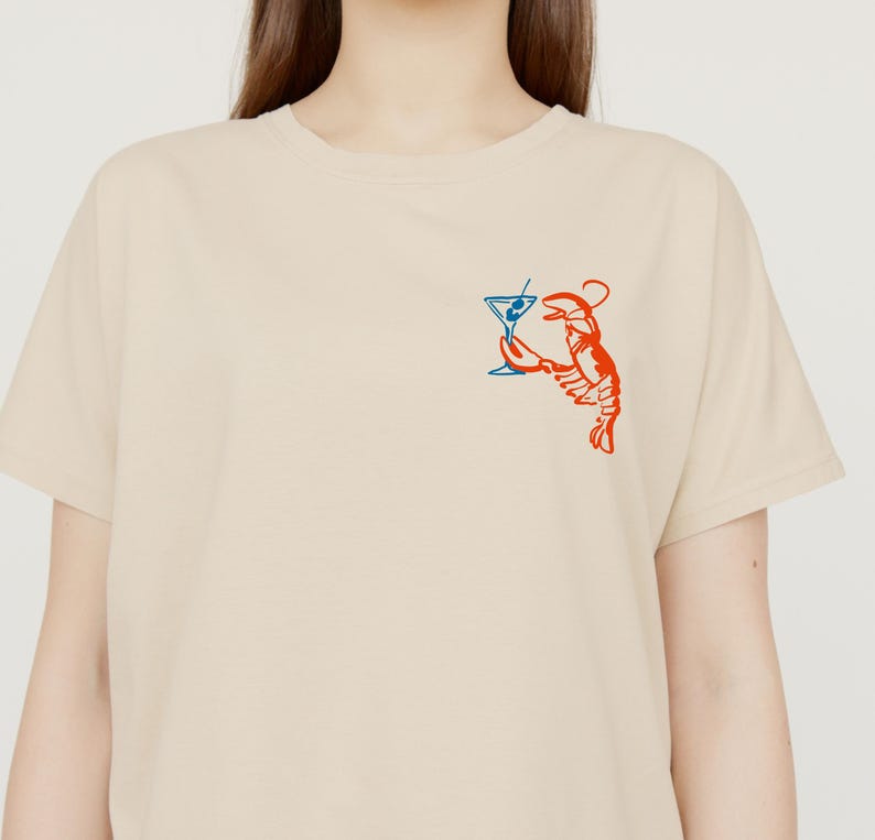 Peut inclure: T-shirt beige avec un motif rouge et bleu d'un homard tenant un verre &agrave; martini. Le homard est dessin&eacute; dans un style fantaisiste, avec le verre &agrave; martini en bleu et le homard en rouge.