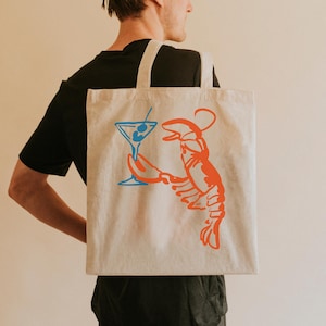 Peut inclure: Sac fourre-tout beige avec une illustration vibrante d'un homard orange tenant un cocktail bleu. Le sac a deux sangles blanches et est fait d'un tissu robuste. Le design est simple et accrocheur, parfait pour un usage quotidien.