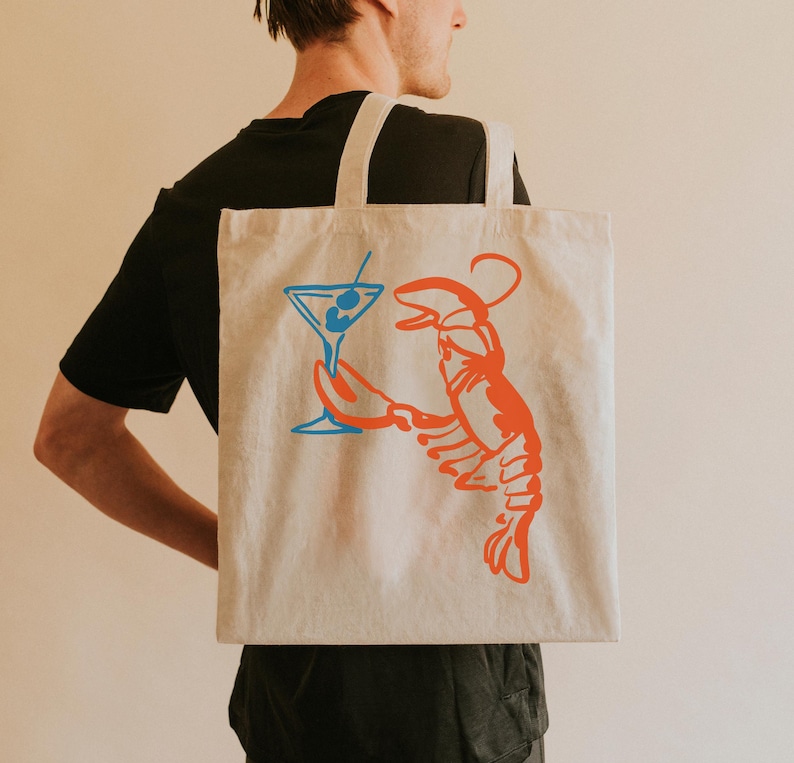 Peut inclure: Sac fourre-tout beige avec une illustration vibrante d'un homard orange tenant un cocktail bleu. Le sac a deux sangles blanches et est fait d'un tissu robuste. Le design est simple et accrocheur, parfait pour un usage quotidien.