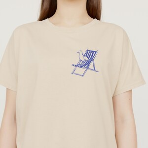 May include: A beige t-shirt featuring a blue illustration of a seagull and deckchair. The graphic is on the left chest. The shirt has a crew neck and short sleeves. The fabric appears to be cotton.