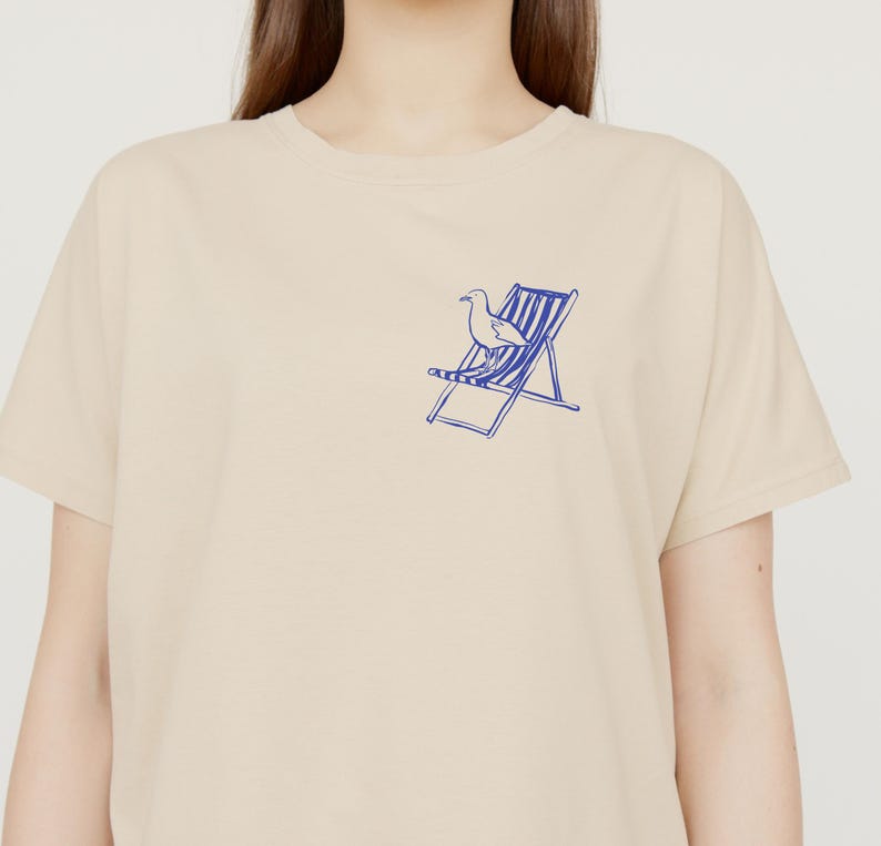 May include: A beige t-shirt featuring a blue illustration of a seagull and deckchair. The graphic is on the left chest. The shirt has a crew neck and short sleeves. The fabric appears to be cotton.