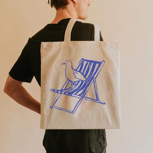 May include: A natural-coloured tote bag featuring a blue outline of a seagull and a deckchair. The bag is carried over a person's shoulder. The design is simple and evokes a seaside theme, perfect for carrying groceries or beach essentials.