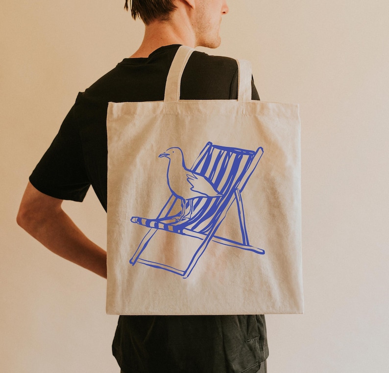 May include: A natural-coloured tote bag featuring a blue outline of a seagull and a deckchair. The bag is carried over a person's shoulder. The design is simple and evokes a seaside theme, perfect for carrying groceries or beach essentials.