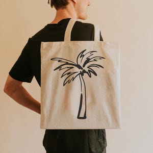 May include: A person is carrying a beige tote bag with a black palm tree design. The bag has a black strap.