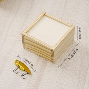 May include: A small, unfinished wooden box, 3.5 inches long, 1.96 inches high, and a gold-colored fishing lure, 2.1 inches long, with two treble hooks. The box and lure are on a light wood-grain surface. The box is rectangular.