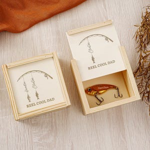May include: Two open wooden boxes with fishing-themed designs. The boxes feature a fishing lure and the text "REEL COOL DAD". The lure is gold-colored with three hooks. The box lids have a fishing rod illustration and the names "Hamilton" and "Clinton".