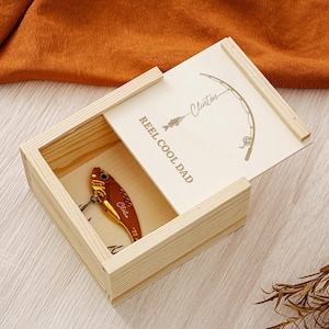 May include: A wooden box with a sliding lid, engraved with a fishing rod and the words "REEL COOL DAD". Inside, a gold and brown fishing lure is visible. The lid is slightly ajar, revealing the contents.