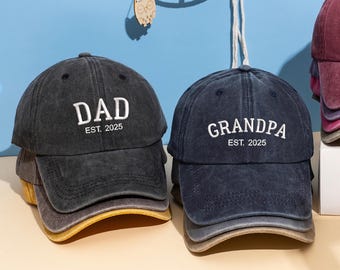 May include: A stack of dark gray and navy blue baseball caps. The dark gray cap has the word "DAD" embroidered in white, with "EST. 2025" below. The navy blue cap says "GRANDPA" with "EST. 2025".