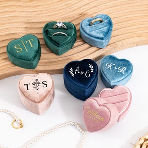 May include: A collection of heart-shaped ring boxes in various colors, including green, blue, and pink. Some boxes feature initials or names in gold or white lettering. The boxes are made of velvet and are designed to hold rings.