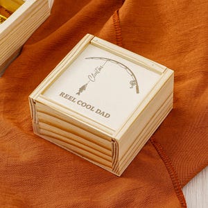 May include: A small, light-colored wooden box with a hinged lid. The lid features an engraved fishing rod design, a fish, and the words "REEL COOL DAD" and the name "Clinton". The box is sitting on a textured orange surface.